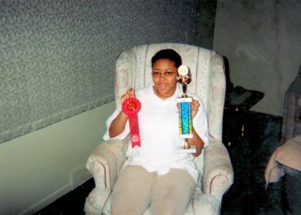 15-year-old Sharee sits in a recliner holding a ribbon in one hand and a trophy in the other.