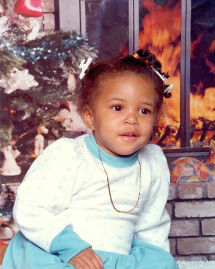 Sharee at one year old, wearing a sweater dress in front of a Christmas tree.