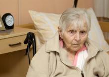 An elderly woman with short hair, sitting propped up in a wheelchair with a pillow.